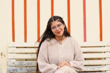 Genç bir kadın bankta poz veriyor, şık örgü kıyafetlerini ve neşeli gülümsemesini canlı çizgilere karşı sergiliyor..