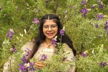 Parlak ve gülümseyen genç bir kadın yemyeşil bir çevrede canlı mor çiçeklerle çevrilidir.