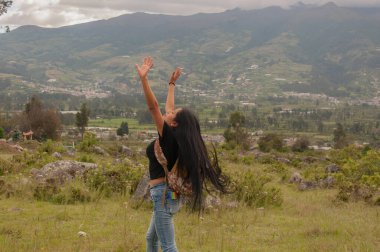 Uzun dalgalı saçları olan bir kadın neşeyle kendini ifade eder nefes kesici güzellikte bir dağ manzarası arasında.