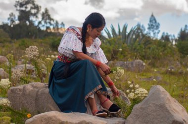 Geleneksel giyinmiş bir kadın kayaların arasında huzur içinde oturup doğanın zarif güzelliğini takdir eder.