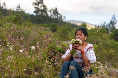 Canlı, geleneksel giysiler giymiş sakin bir kadın yemyeşil bir manzarada çiçeklerin güzelliğinden zevk alır.