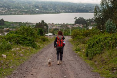 Arka planda çarpıcı bir göl olan köpeğini toprak yolda mutlu bir şekilde gezdiren bir kadının huzurlu görüntüsü.