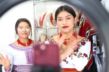 Geleneksel nakışlı kıyafetler ve kırmızı kolyeler takan iki genç yerli kadın, kültürlerini ve modalarını internette tanıtmak için tripoda akıllı telefonlu bir video kaydediyorlar.