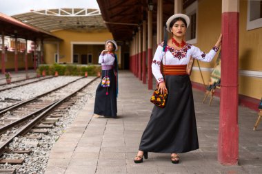 Geleneksel Andean kıyafetleri ve şapkaları giyen iki yerli otavalo kadın, zengin kültür mirasını ve moda anlayışlarını sergilemek için tren istasyonunda poz veriyorlar.