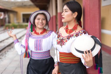 Geleneksel Andean kıyafetleri ve şapkaları giyen iki gülümseyen yerli otavalo kadın tren istasyonunda poz veriyor, zengin kültürel miraslarını renkli işlemeli bluzlar ve kemerlerle sergiliyorlar.