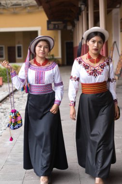 İki gülümseyen yerli otavalo kadın, geleneksel el yapımı bluzlarını ve siyah eteklerini giyerek bir koloni caddesinde yürüyorlar. Ekvador ve dekan kültürünü temsil ediyorlar.