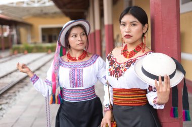 Geleneksel ve nakışlı bluzlar ve renkli kemerler giyen iki yerli riobamba kadını bir tren istasyonunda poz veriyor, zengin kültürel mirasını ve eşsiz moda anlayışlarını sergiliyorlar.