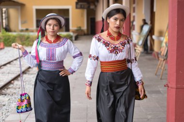 Geleneksel ve işlemeli bluz ve siyah etek giyen iki genç otavalo kadın bir tren istasyonunda yürüyor ve zengin kültürel miraslarını sergiliyorlar.