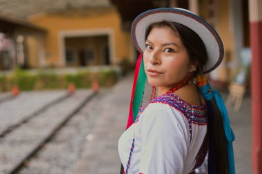 Beyaz şapkalı ve kurdeleli geleneksel renkli elbiseli yerli bir kadın bir tren istasyonunda kültürel mirasını sergilemek için poz veriyor.