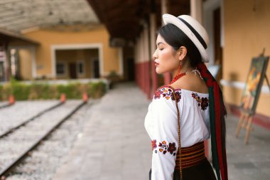 Yerli kadın bir tren istasyonunda zarif bir şekilde duruyor, geleneksel giysilerini karmaşık nakış ve canlı aksesuarlarla sergiliyor. Sahne kültür mirası ve modaya vurgu yapıyor