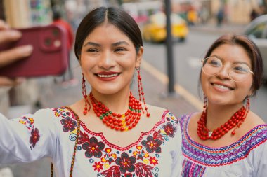 Canlı geleneksel kıyafetler giymiş ve boncuklu kolyelerle süslenmiş iki neşeli yerli kadın hareketli bir caddede neşeli bir selfie çekiyorlar.