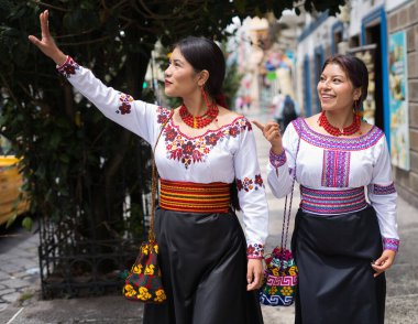 Geleneksel ve nakışlı bluzlar ve renkli kemerler giyen iki gülümseyen yerli otavalo kadın caddede yürüyor ve küçük bir ekvador şehrinde taksi çağırıyorlar.