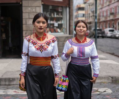 Geleneksel otavalo kıyafetleri ve mücevherleri giyen iki ekvatorlu kadın, canlı kültürlerini ve mirasını sergilemek için kaldırım taşından bir sokakta yürüyorlar.