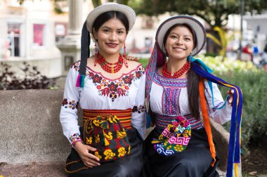 Geleneksel riobamba giysileri ve mücevher giyen iki ekvator kadın zengin kültür mirasını temsil eden bir parkta taş bir bankta poz veriyorlar.
