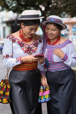 İki gülümseyen yerli riobamba kadını, canlı geleneksel giyinmiş ve şapka takmış, iletişim ve bağlantı için akıllı telefon kullanırken bir şehir caddesinde birlikte yürüyorlar.