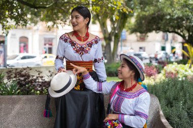 Geleneksel Andean giysileri giyen iki yerli kadın parkta bir bankta oturmuş, bir şapkayı işaret ediyor ve kültürlerini ve mirasını tartışıyorlar.