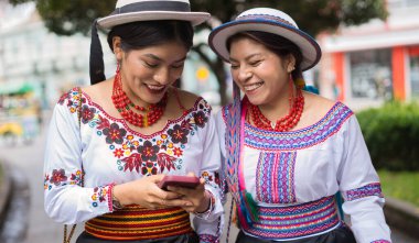 Geleneksel işlemeli bluz ve şapkalar giyen, gülümseyen ve akıllı bir telefonla meşgul olan iki yerli kadın. Canlı kıyafetleri kültürel mirası ve modern bağlantıyı yansıtıyor.