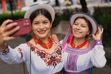 Geleneksel andean kıyafetleri ve şapkalar giyen iki mutlu yerli kadın akıllı telefonlarıyla selfie çekiyor, gülümsüyor ve kucaklıyor, kültürlerini ve mirasını kutluyorlar.