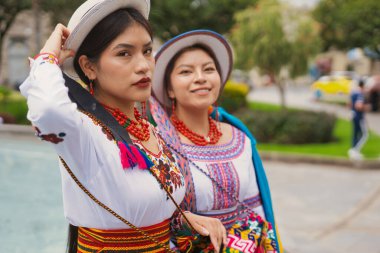 Geleneksel Andean giysileri ve şapkalar giyen iki gülümseyen yerli otavalo kadın, zengin kültürel miraslarını sergilemek için parkta poz veriyorlar.