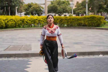 Genç otavalo kadın gururla canlı renkler sergiliyor ve kültürel gururun sembolü olarak bir şehir parkında yürürken geleneksel ve dekan kıyafetlerinin karmaşık nakışlarını sergiliyor.