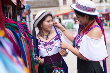 İki yerli kadın, zengin kültürel mirasını kutlamak için canlı ve renkli bir pazar yerinde geleneksel kıyafet ve aksesuar denerken gülümsüyor.