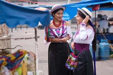 Geleneksel Andean kıyafetleri ve şapkalar giyen iki gülümseyen yerli riobamba kadını, Ekvador 'daki otavalo pazarında bir meyve tezgahının yanında konuşuyorlar.