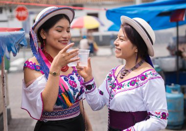 Canlı, nakışlı geleneksel giysiler ve şapkalar giyen iki yerli kadın, hareketli bir pazar yerinde plastik bardaklardan gelen ferahlatıcı bir içkiyi paylaşarak birlikte bir anın tadını çıkarıyorlar.