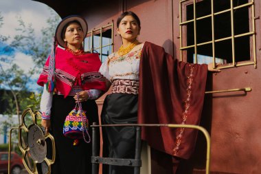İki yerli otavalo modeli, geleneksel Andean giysileri, mücevherleri ve aksesuarlarıyla eski bir tren vagonunun yanında poz veriyor.