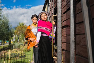 Geleneksel And giysileri ve aksesuarları giyen iki yerli otavalo kadın, Ekvador 'da güneşli bir günde eski bir ahşap tren vagonunun yanında poz veriyorlar.