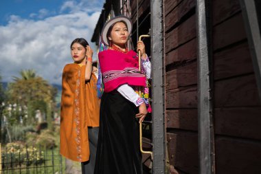 Geleneksel Andean giysileri giyen iki ekvator kadını, tarihi ve kültürel gururu temsil eden eski bir ahşap tren vagonunun yanında poz veriyorlar.