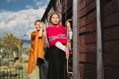 Geleneksel Andean giysileri giyen iki yerli manken, canlı kıyafetlerini ve kültürel miraslarını sergileyen metal detaylarla dolu ahşap bir vagonun yanında poz veriyorlar.