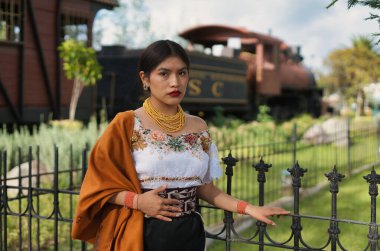 Geleneksel And giysileri ve altın mücevher giyen genç bir kadın, kültürel mirasını gururla ve zarafetle temsil eden bir parkta, eski bir buhar treninin yanında poz veriyor.