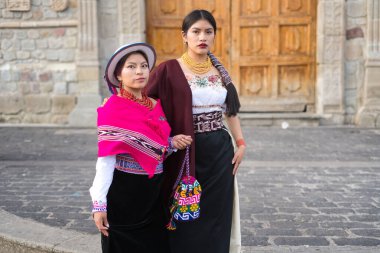 Geleneksel Andean giysileri giyen iki ekvatorlu kadın, kültürlerinin zengin tekstil mirasını sergilemek için bir koloni kilisesi önünde birlikte poz veriyorlar.