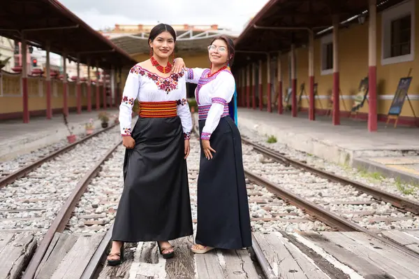 İki genç otavalo yerlisi kadın tren istasyonunun raylarında duruyorlar, geleneksel kıyafetlerini ve aksesuarlarını giyip birlikte poz veriyorlar.