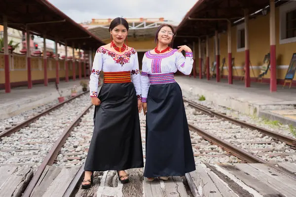 Geleneksel yerli kıyafetlerini gururla sergileyen iki kadın tarihi bir tren istasyonunda demiryolu raylarında dikiliyorlar. Canlı kıyafetleri kültürel mirası ve modern moda birleşimini vurguluyor.
