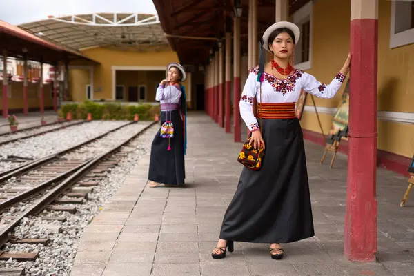 Geleneksel Andean kıyafetleri ve şapkaları giyen iki yerli otavalo kadın, zengin kültür mirasını ve moda anlayışlarını sergilemek için tren istasyonunda poz veriyorlar.
