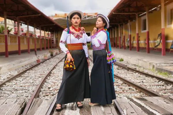 Geleneksel Andean kıyafetleri ve şapkaları giyen iki yerli otavalo kadın, zengin kültür mirası ve modalarını sergileyerek tren raylarında poz veriyorlar.