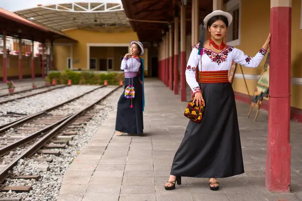 İki genç otavalo yerlisi kadın tren istasyonunda poz veriyorlar, geleneksel ve renkli bluzlar, renkli kemerler ve uzun siyah etekler giyiyorlar.