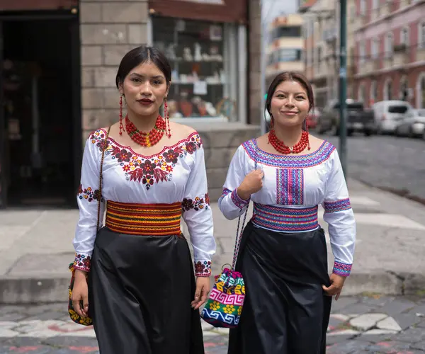 Geleneksel otavalo kıyafetleri ve mücevherleri giyen iki ekvatorlu kadın, canlı kültürlerini ve mirasını sergilemek için kaldırım taşından bir sokakta yürüyorlar.
