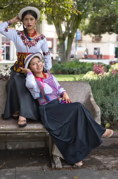 Canlı bir parkta, taştan bir bankta otururken, geleneksel yerli kıyafetli iki kadın renkli vitrinler sergiliyorlar. Kıyafetleri kültürel mirası ve modern tarzı yansıtıyor.