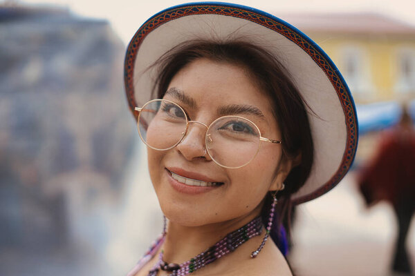 Young indigenous woman wearing traditional hat, glasses, earrings and necklace, representing her rich cultural heritage and embodying modern indigenous fashion with a cheerful smile