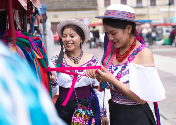 Geleneksel andean kıyafetleri ve şapkalar giyen iki gülümseyen yerli kadın açık hava pazarında renkli kurdeleler seçiyor ve birlikte alışveriş deneyimlerinin tadını çıkarıyorlar.
