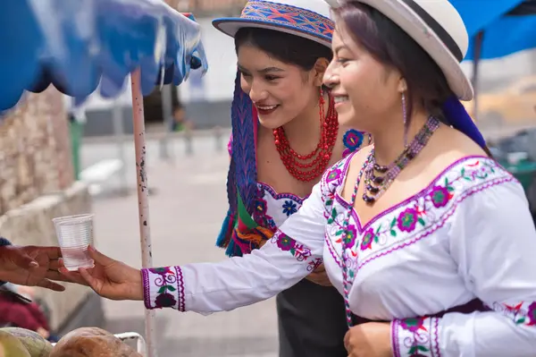 Geleneksel kıyafetler ve şapkalar giyen iki yerli kadın, canlı kültürlerini ve toplumsal ilişkilerini sergilemek için açık hava pazarındaki bir satıcıdan bir bardak taze meyve suyu alıyorlar.