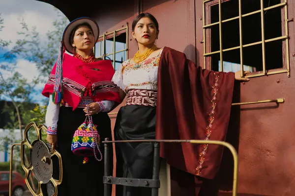 İki yerli otavalo modeli, geleneksel Andean giysileri, mücevherleri ve aksesuarlarıyla eski bir tren vagonunun yanında poz veriyor.