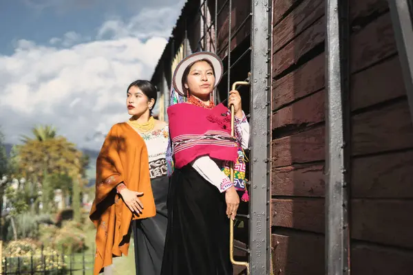 İki genç yerli kadın, canlı geleneksel Andean kıyafetleri ve aksesuarları giyip, eski bir tren vagonunun yanında poz vererek kültürel mirasını ve çağdaş modasını kutluyorlar.