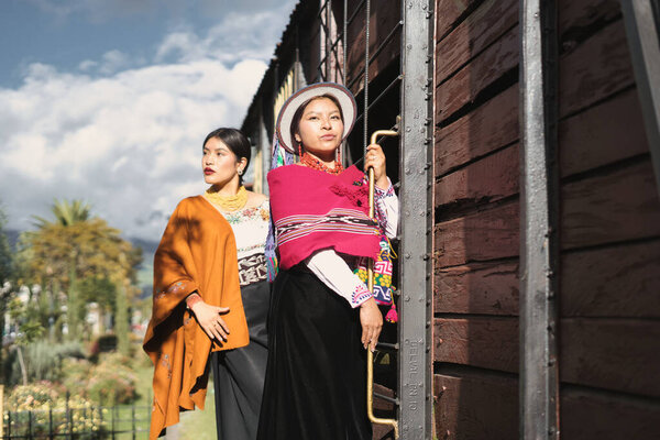 Two young indigenous women, dressed in vibrant traditional andean clothing and accessories, posing beside a vintage train wagon, celebrating their cultural heritage and contemporary fashion