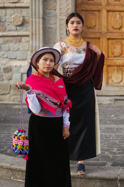 Two ecuadorian women proudly displaying vibrant traditional attire, featuring embroidered blouses, long skirts, and handcrafted accessories, stand before a historic stone building