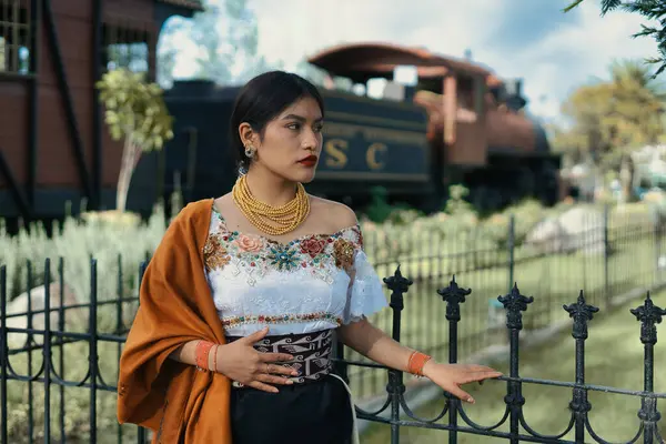 Geleneksel kıyafetler ve altın mücevherler giyen yerli bir kadın, arkasında bulanık bir buhar treniyle siyah metal bir çitin yanında poz veriyor ve kültürel mirasını ve gururunu sergiliyor.