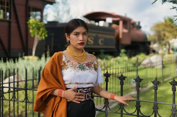 Geleneksel And giysileri ve altın mücevher giyen genç bir kadın, kültürel mirasını gururla ve zarafetle temsil eden bir parkta, eski bir buhar treninin yanında poz veriyor.