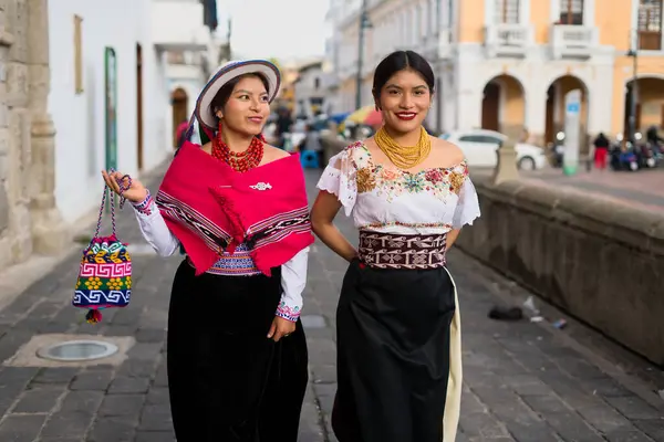 Geleneksel Andean giysileri ve mücevherleri giyen iki gülümseyen yerli otavalo kadın, zengin kültürel miraslarını sergilemek için Ekvador 'daki bir şehirde yürüyorlar.